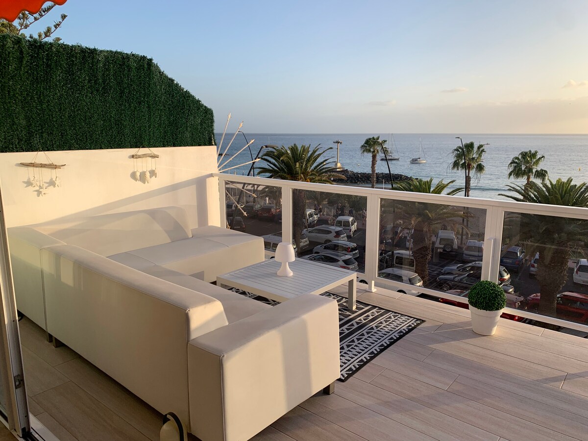 An outdoor terrace features modern lounge seating with clean lines, complemented by a small coffee table. Expansive ocean views are visible beyond the railing, with palm trees and parked cars lining the beach. Soft evening light enhances the tranquil atmosphere.