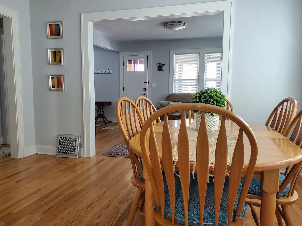 A dining area is showcased with a wooden table surrounded by six chairs, highlighting a welcoming space for meals. Natural light filters through the open doorway, illuminating the adjacent living room, which features a comfortable sofa and a front door with glass panes.