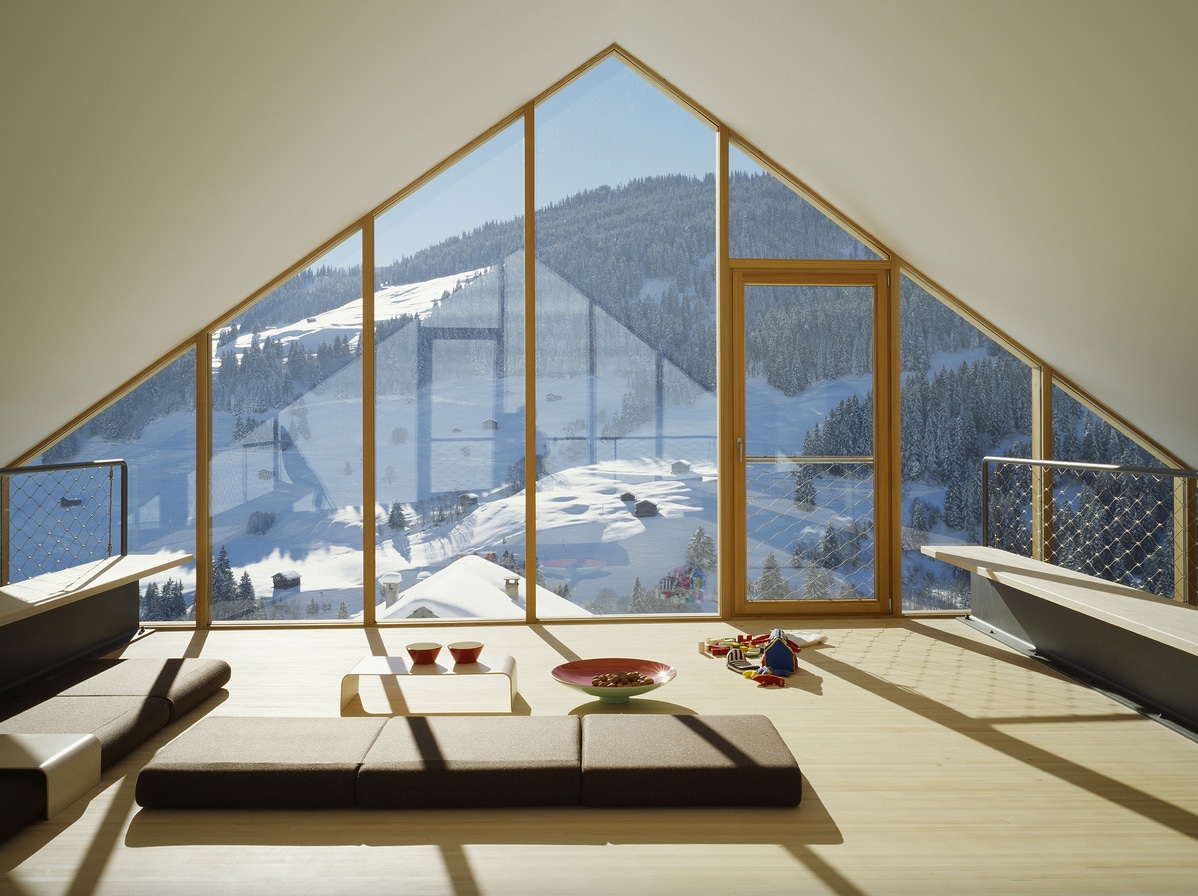 A spacious room with a sloped ceiling features large windows that provide expansive views of the snowy landscape outside. Light wood flooring complements a low seating arrangement, and a coffee table is positioned in the center. Bright colors from decorative objects enhance the inviting atmosphere.