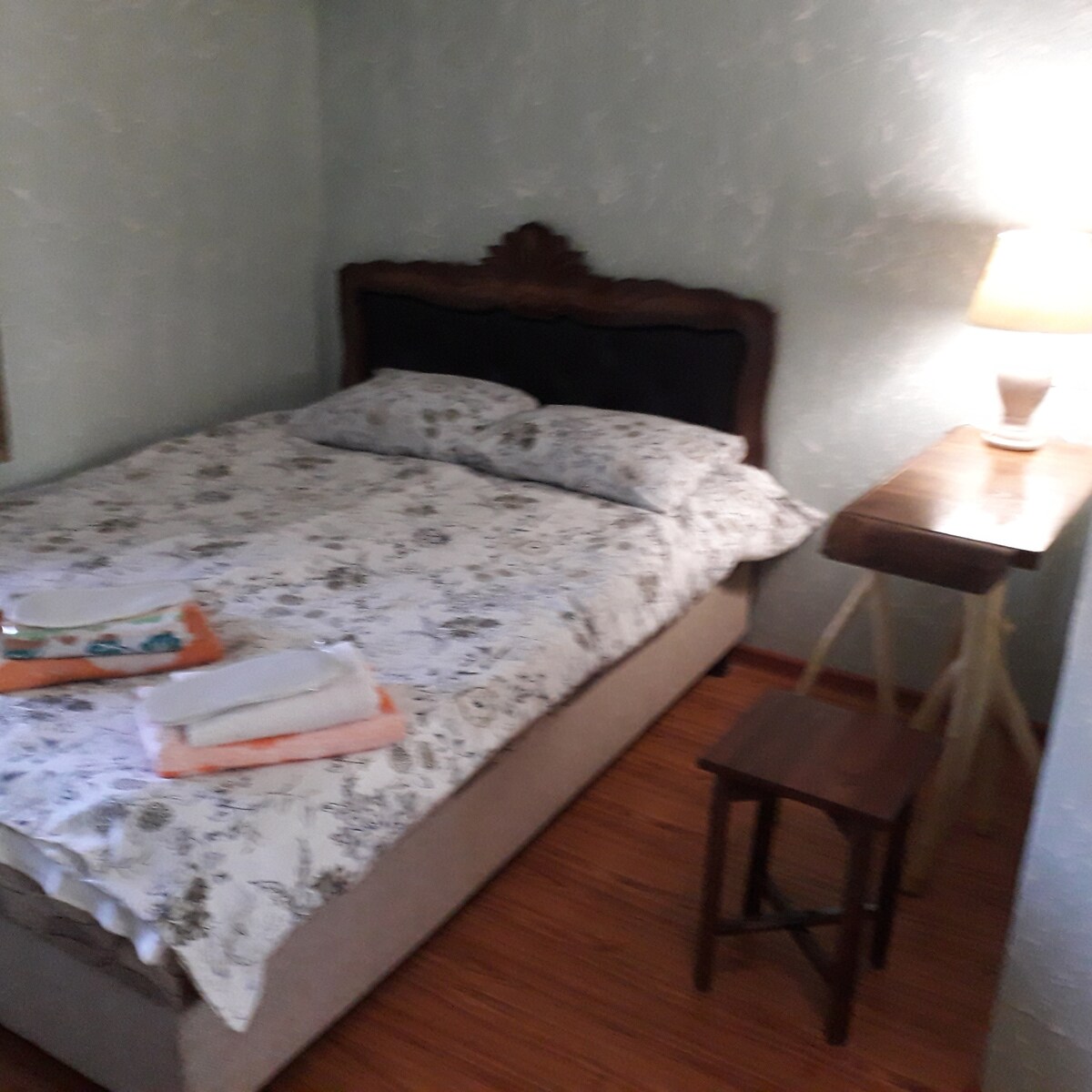 A cozy sleeping area features a spacious bed with a floral-patterned duvet cover. Neatly folded towels are arranged on a side table next to the bed, which is complemented by a simple lamp providing soft illumination. A small wooden stool adds functional charm to the room.