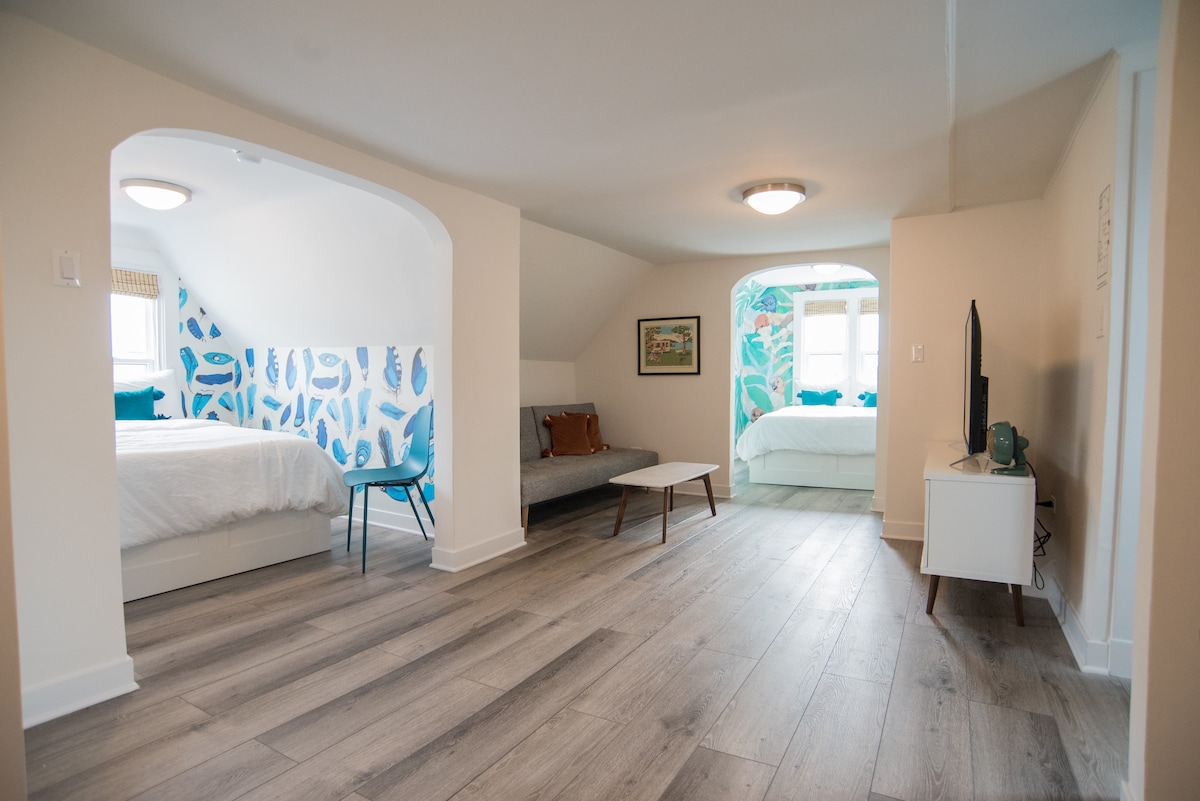 A spacious living area features two semi-private bedrooms, with soft natural light illuminating the light wood flooring. Colorful wall art with leaf patterns adds a touch of vibrancy. A small couch and a coffee table are arranged in the center of the room, alongside a television mounted on a white cabinet.