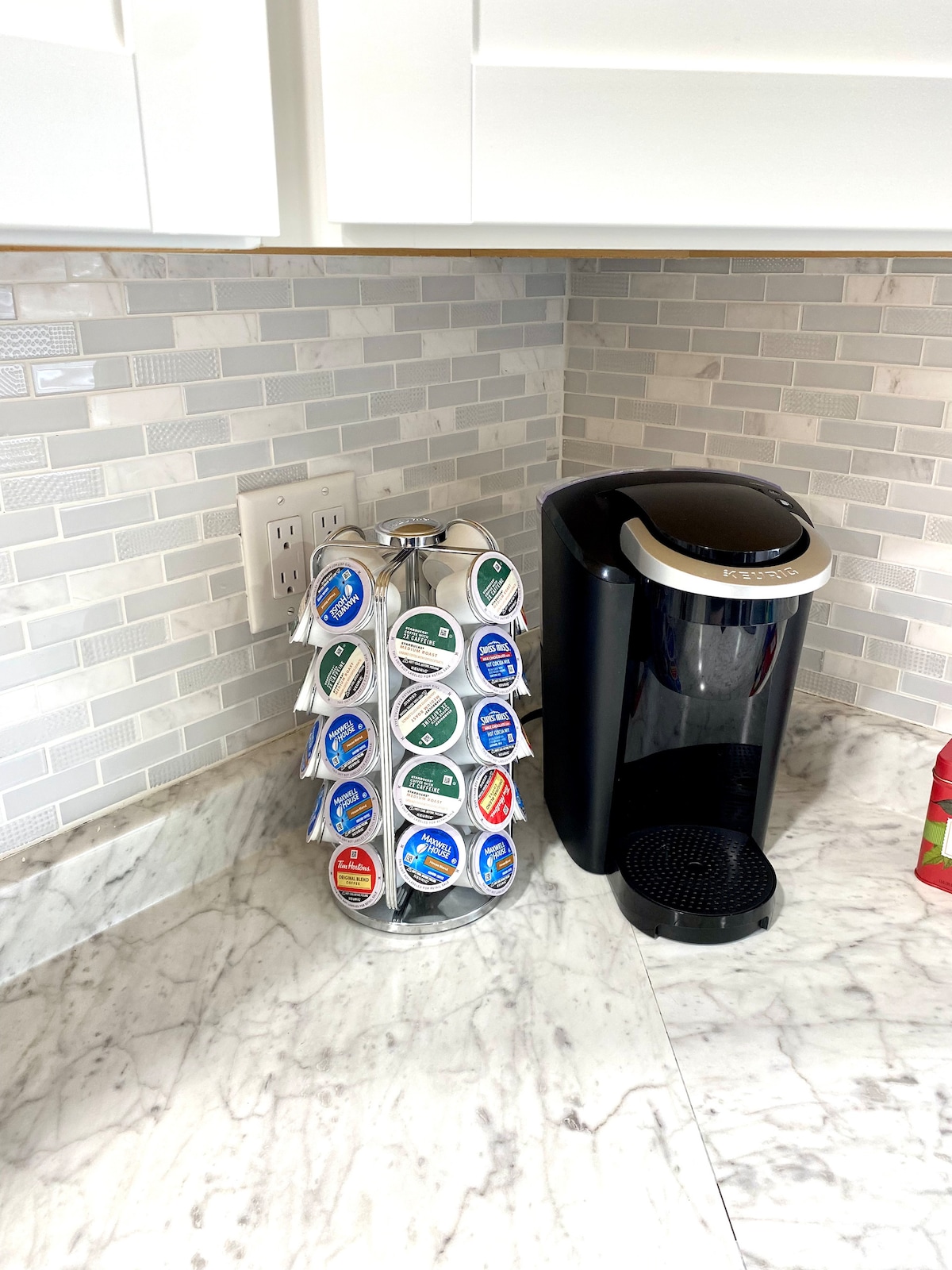 A coffee station is displayed featuring a modern coffee maker next to a carousel of assorted coffee pods. The counter is topped with light-colored marble, while a tiled backsplash in soft grey tones adds a subtle texture to the kitchen space.