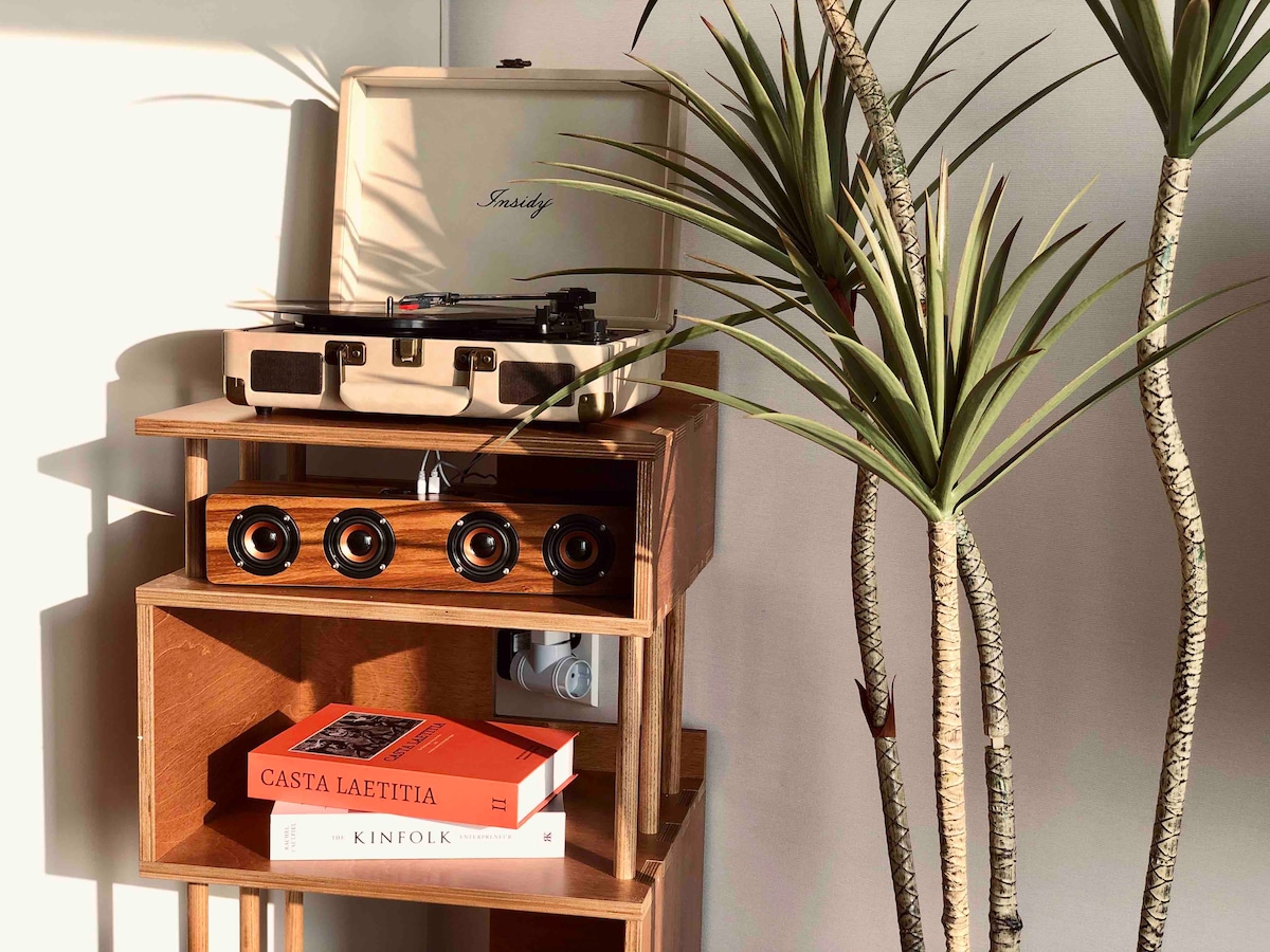 A stylish wooden shelf holds a turntable atop, surrounded by potted plants. Below, books are neatly arranged, with a patterned book cover visible. Natural light highlights the texture of the wood and the green leaves, creating a serene and inviting corner.
