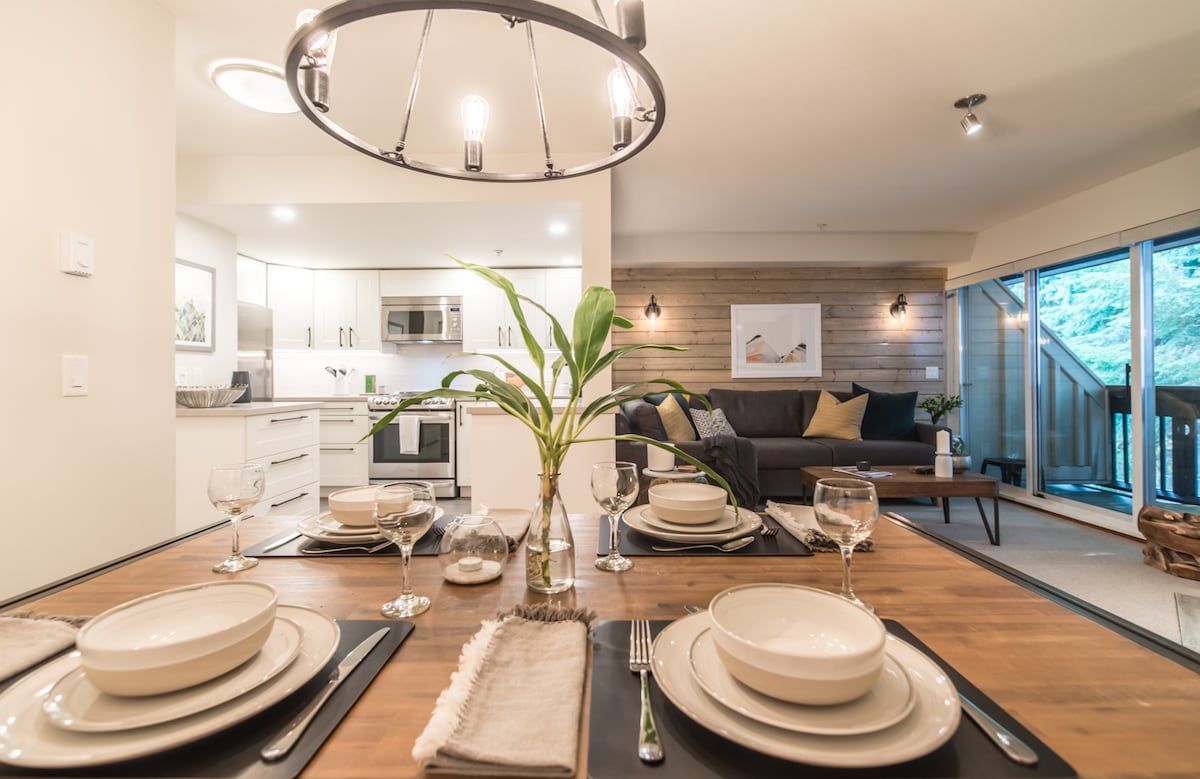 A dining area features a wooden table set with dishware, glasses, and cutlery. In the background, a modern kitchen is visible, equipped with stainless steel appliances and ample cabinetry. A cozy living room is seen through large glass doors, showcasing a comfortable sofa and natural light.