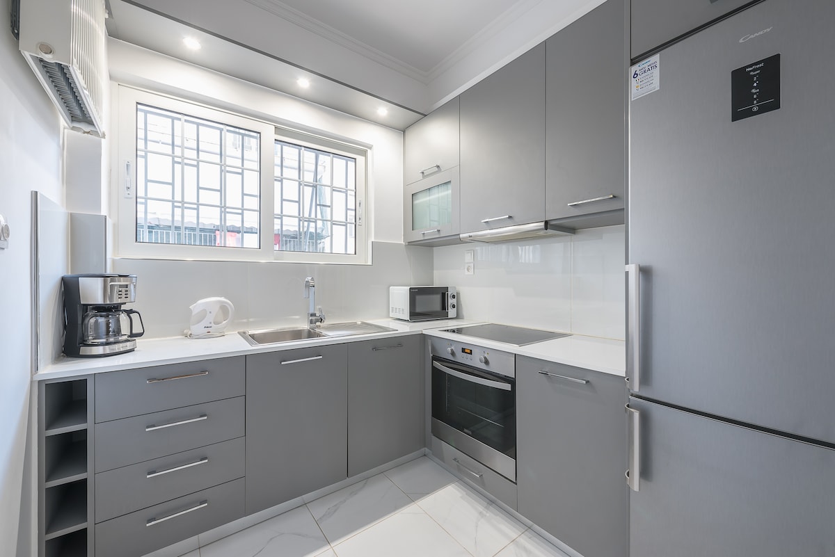 A modern kitchen is arranged with sleek gray cabinets and white countertops. Essential appliances include a refrigerator, microwave, and coffee maker. Natural light enters through a window above the sink, while additional storage is provided by lower cabinets and drawers.