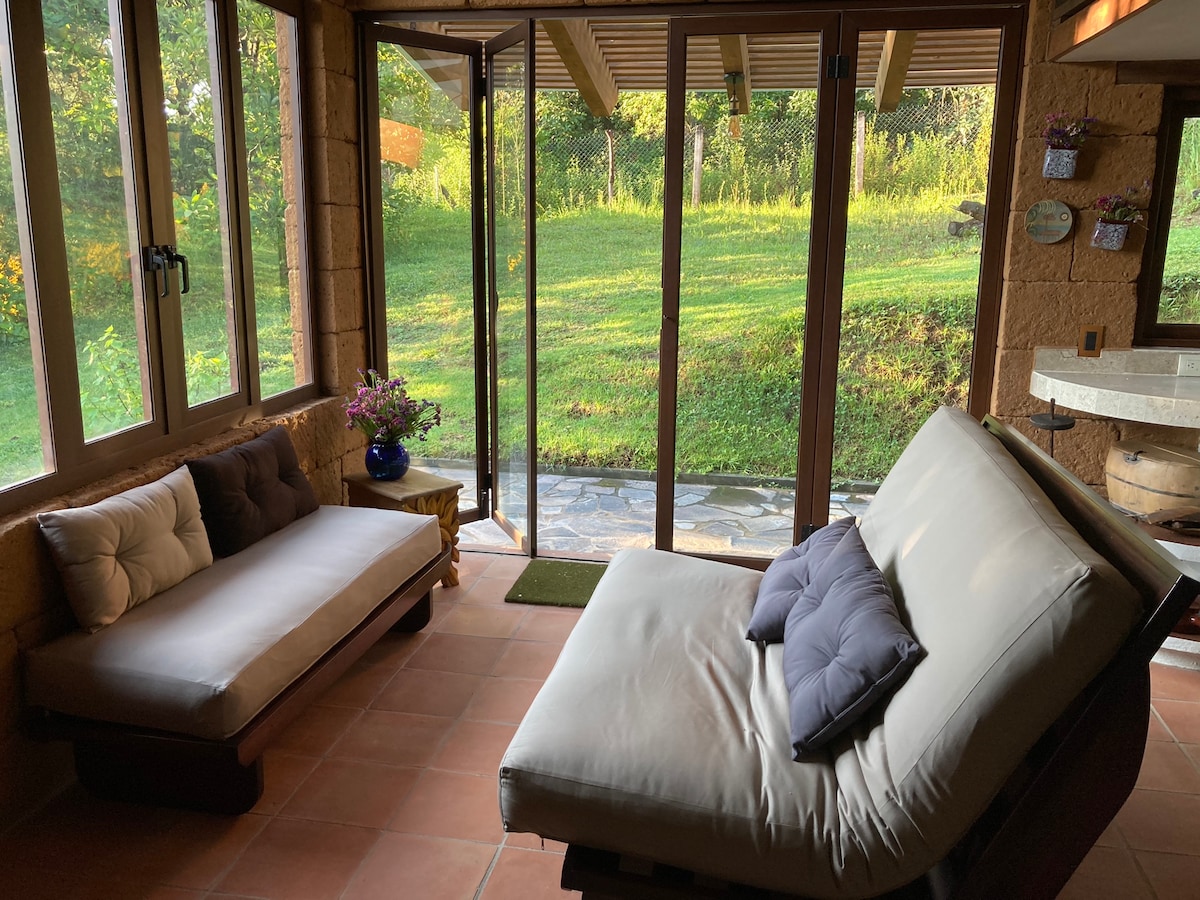 An inviting seating area is framed by large glass doors that open to a green landscape. A spacious sofa with soft cushions creates a comfortable space for relaxation, while natural light illuminates the room, enhancing the connection between indoor and outdoor environments.