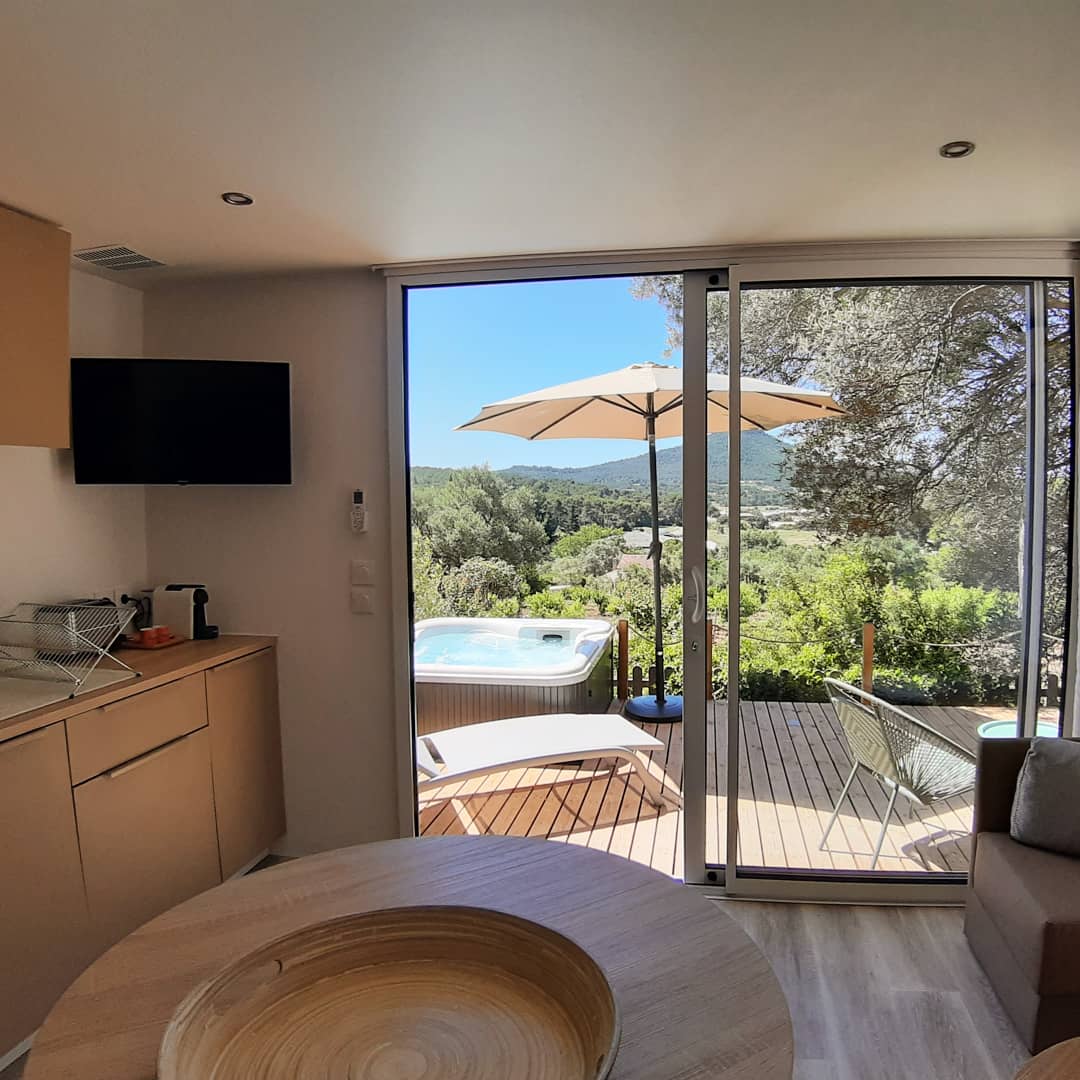 A modern interior showcases a compact kitchenette and a round wooden table. Floor-to-ceiling glass doors provide a view of the lush outdoor landscape and a private jacuzzi on the terrace, complemented by an umbrella and lounging chairs.