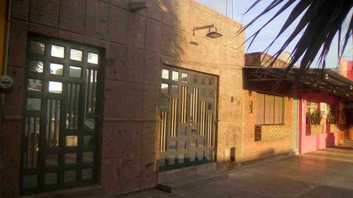 A two-story building is presented, featuring textured stone walls and large windows with vertical elements. A covered entrance is visible, providing access to the interior. The exterior is adorned with greenery, offering a shaded area near the entrance.