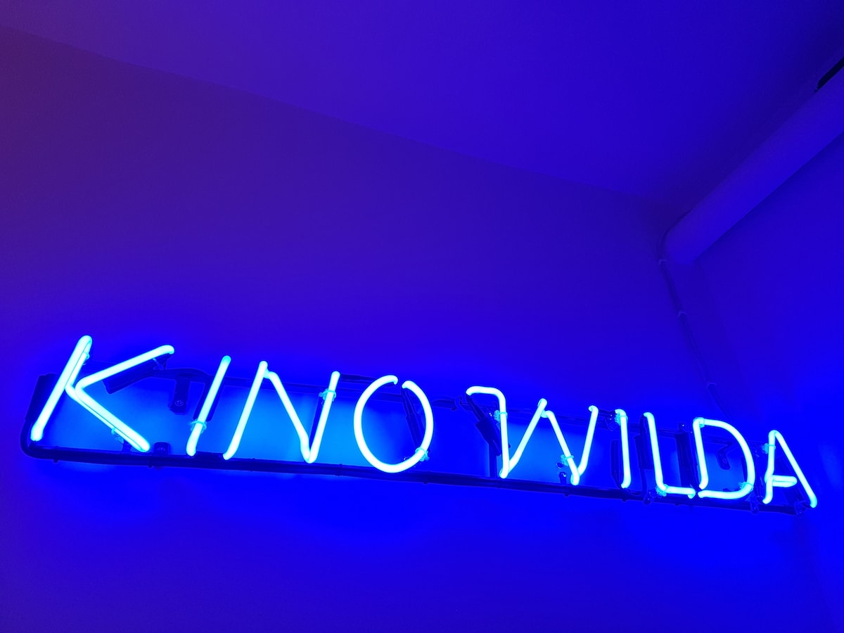 A neon sign displaying the words 'KINO WILDA' in bright blue light adorns the wall, adding a distinctive touch. The vibrant glow contrasts against the neutral wall, creating a visually striking focal point that reflects the apartment's cinematic heritage.