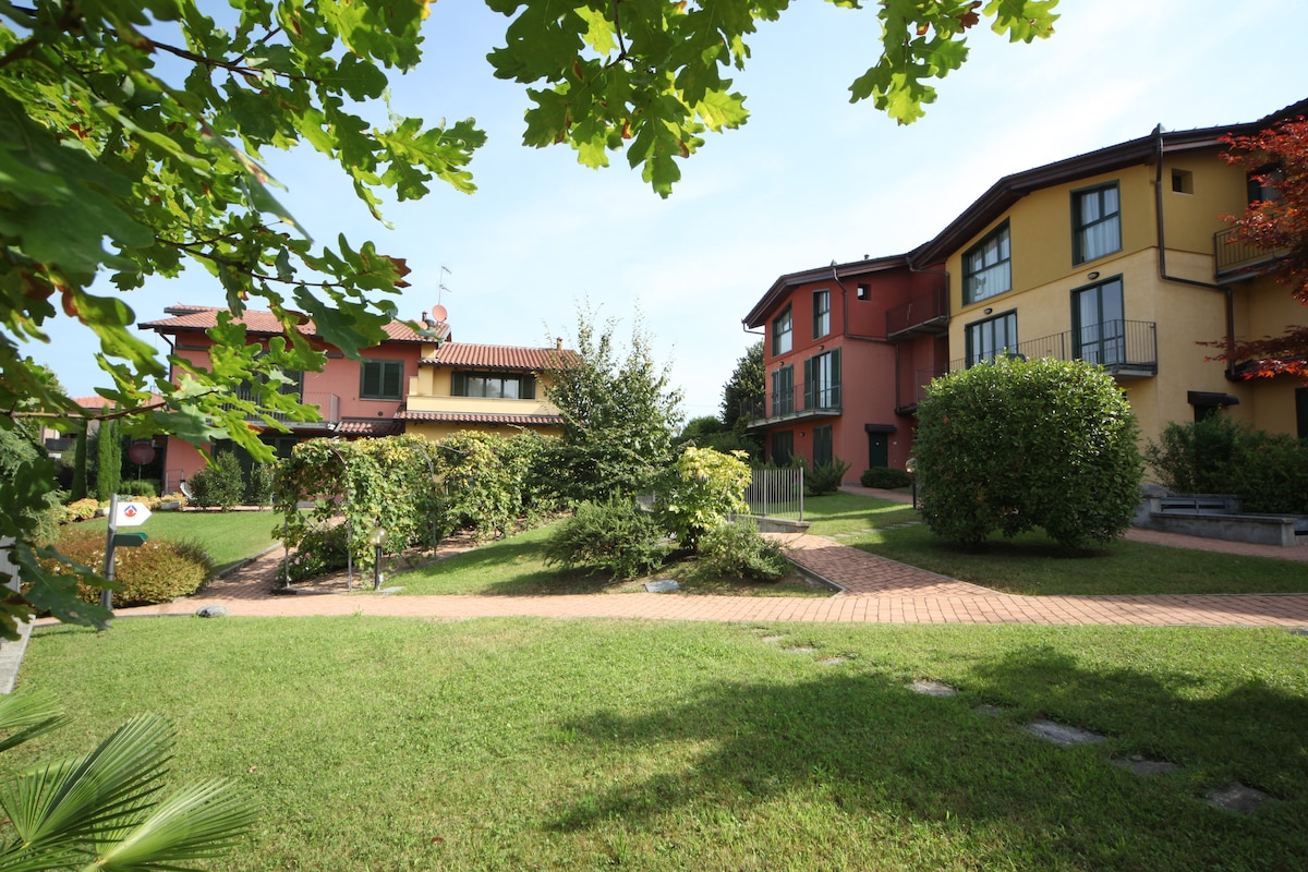 The image displays a serene garden area surrounded by several residential buildings. Lush green lawns and trees are visible, with pathways connecting the structures. The buildings showcase a variety of colors, and balconies can be seen on some units, enhancing the inviting atmosphere.