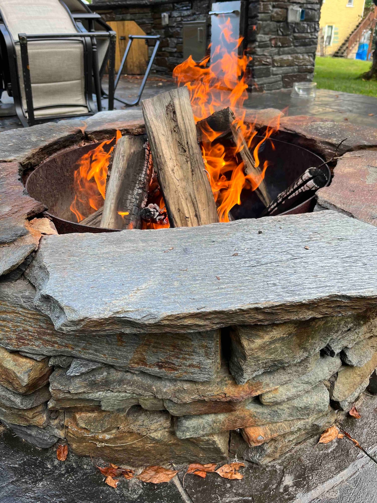 A stone fire pit is shown with stacked stones and a flat stone surface. Bright flames rise from logs placed inside, creating a warm focal point. The area surrounding the fire pit is partially visible, suggesting a social outdoor space.