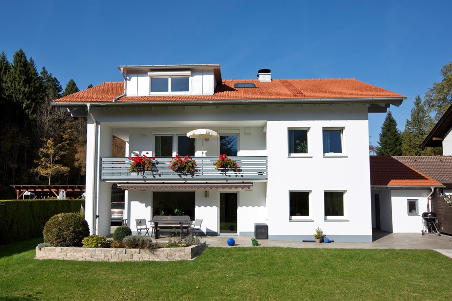 A spacious two-story building is presented, featuring a red-tiled roof and white exterior. A balcony with vibrant flowers overlooks a green lawn, while outdoor seating is arranged below. Large windows allow natural light to fill the interior, surrounded by wooded areas.