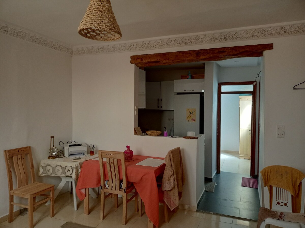An open living area is visible, featuring a small dining table with orange tablecloths and several wooden chairs. A kitchen is partially seen in the background, with modern appliances. Natural light brightens the space, complemented by traditional decor elements like a woven light fixture.