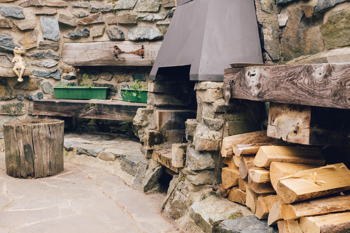 An outdoor grilling area features a stone structure with a metal chimney. Wooden benches provide seating alongside planters. A rough-hewn log serves as a table, while neatly stacked firewood is stored nearby, contributing to a rustic and functional atmosphere.