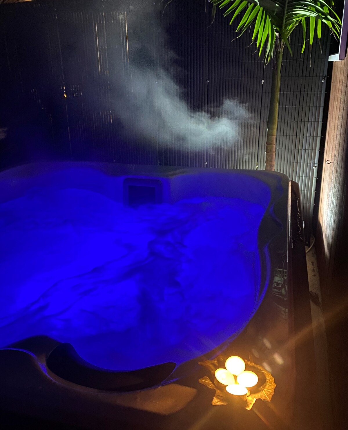 A private hot spa is visible, featuring calming blue water and soft steam rising from the surface. A small cluster of candles gently illuminates the area, adding warmth to the atmosphere. Lush greenery can be seen in the background, enhancing the serenity of the space.