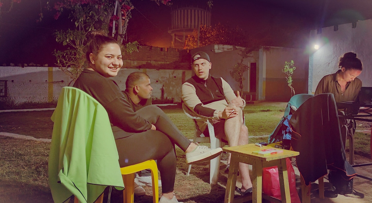 The outdoor seating area features four plastic chairs arranged in a relaxed manner on a grassy surface. A small table is positioned at the center, with several items visible on its surface. Soft lighting enhances the ambiance, creating a welcoming environment for gatherings.
