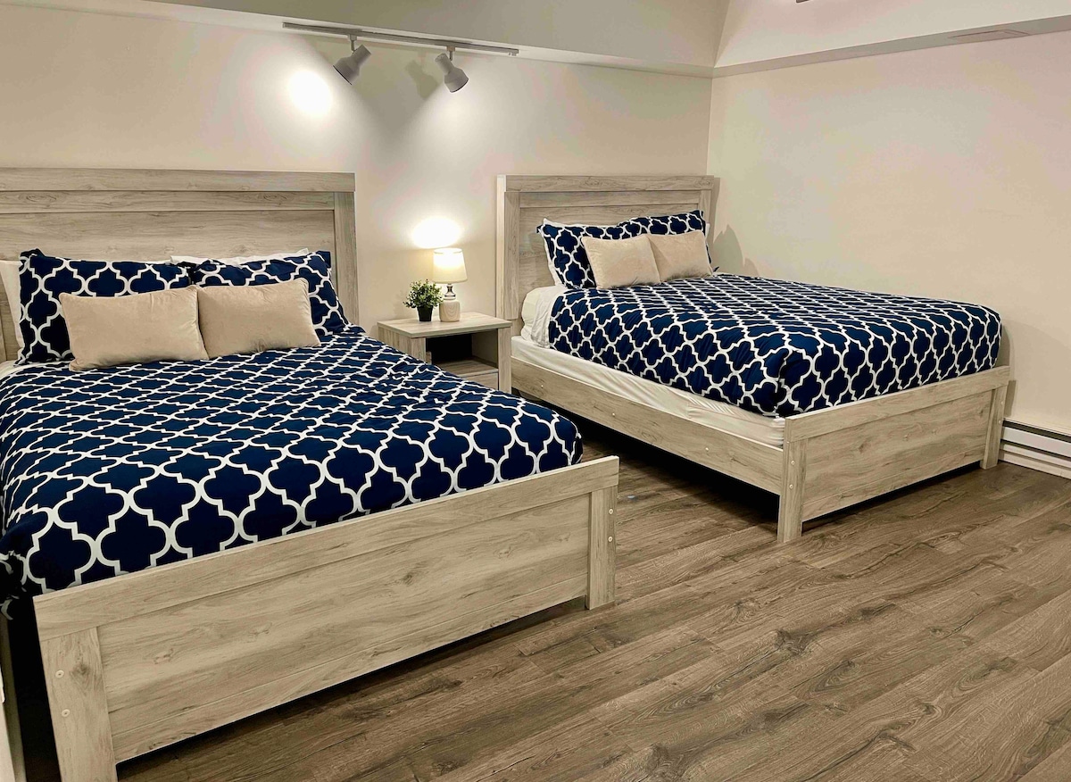 A serene bedroom features two queen-sized beds with blue patterned comforters and neutral accent pillows. A small bedside table with a lamp and a decorative plant are positioned between the beds, while the light wood flooring adds a warm touch to the space.
