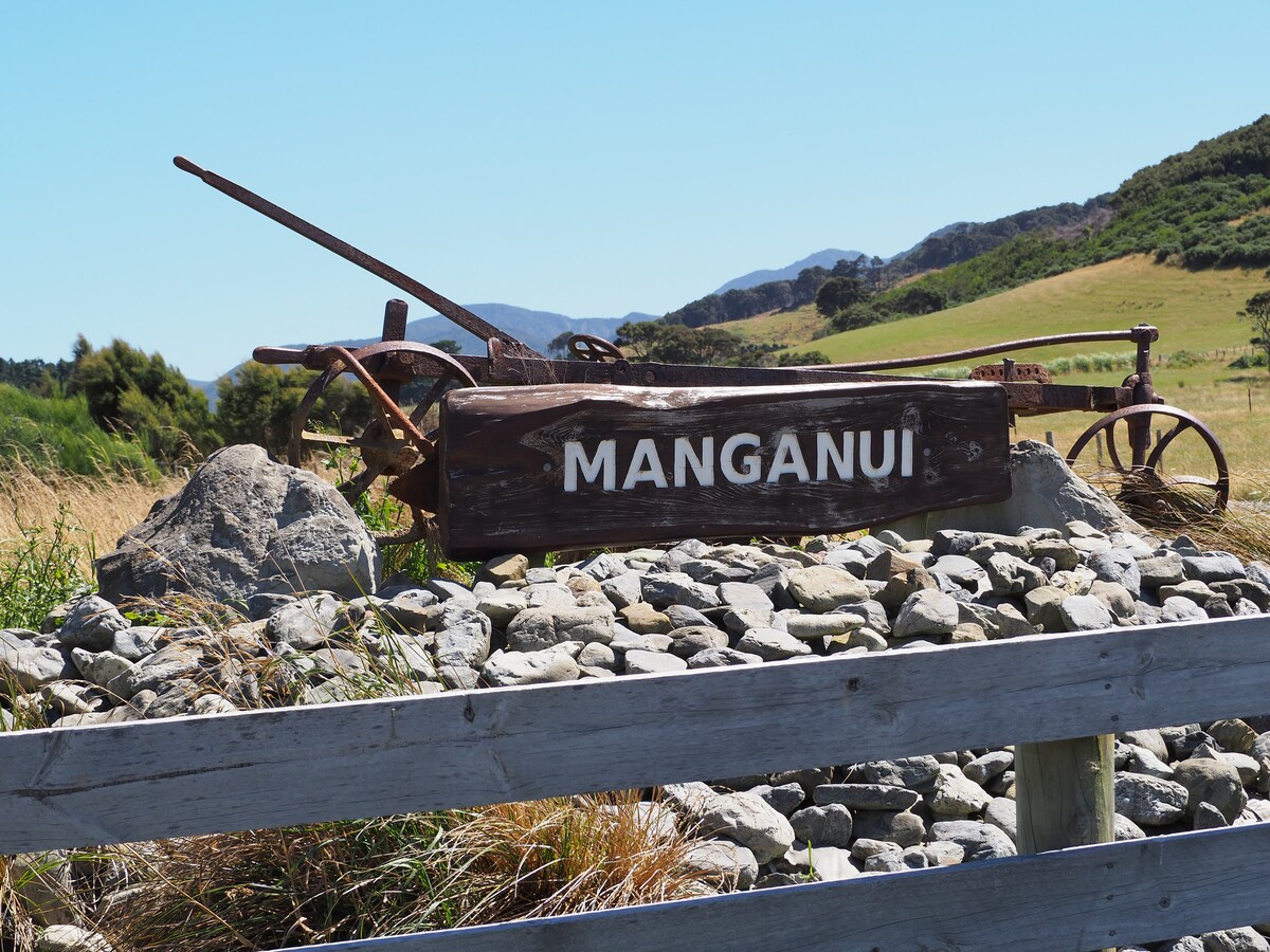 A weathered wooden sign labeled 'Manganui' is positioned prominently among flattened stones, with the backdrop of rolling hills and greenery. An old farming implement is partially visible, complementing the rural landscape.
