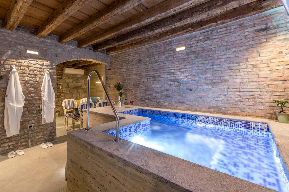 An indoor pool is set against exposed brick walls, featuring a tiled water surface in shades of blue. Two white bathrobes are draped over a nearby railing. The area is illuminated by understated lighting, enhancing the serene atmosphere.