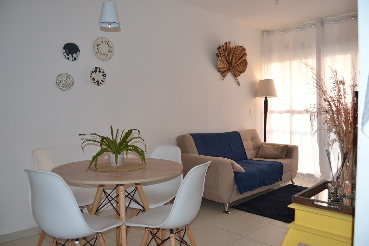 A bright living area features a round wooden dining table surrounded by white chairs. A light beige sofa is positioned nearby, with a decorative wall display showcasing natural materials. Sunlight streams through sheer curtains, illuminating the clean, minimalist decor.