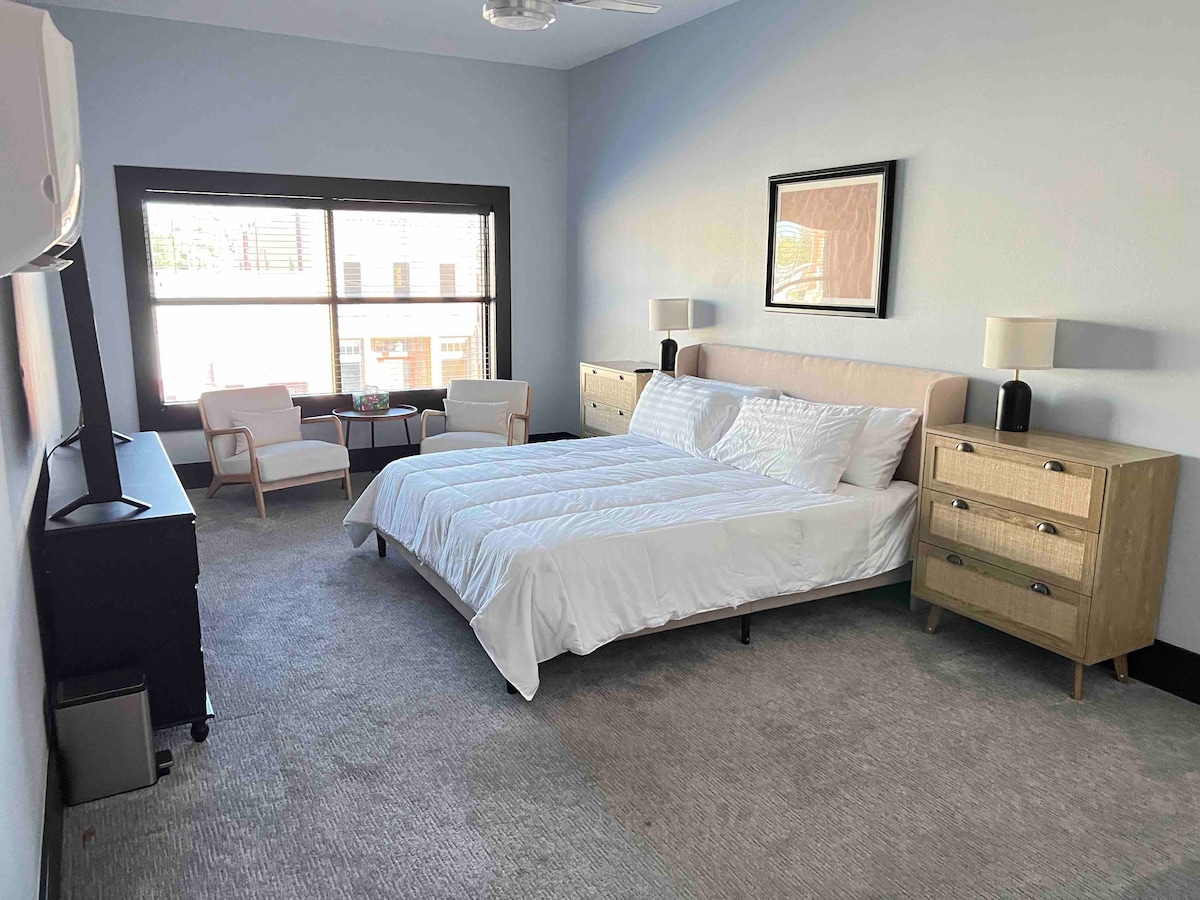 The hotel room features two cozy armchairs beside a small table, positioned near a large window that allows natural light to fill the space. A spacious bed is complemented by a light duvet, while a dresser and TV occupy the opposite wall.