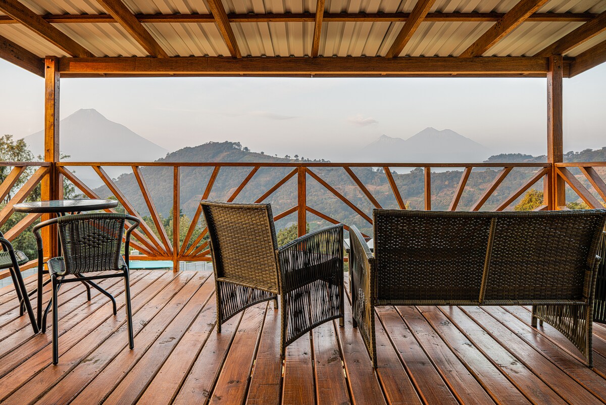 A wooden terrace features woven chairs and a small table, offering expansive views of the surrounding mountains. The structure's design provides an inviting space to appreciate the natural beauty of the landscape, exposed under a soft morning light.
