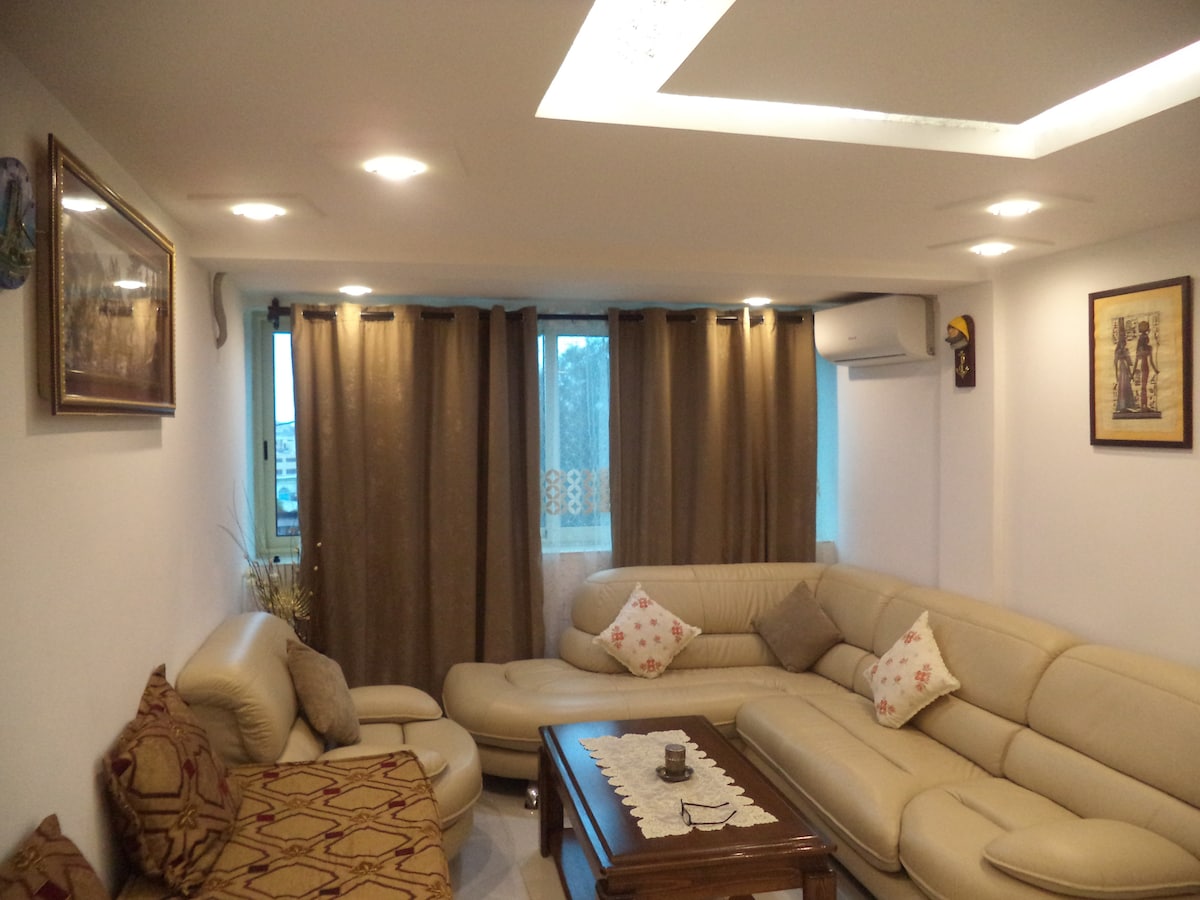 A spacious living area is furnished with a light-colored sectional sofa and a coordinating armchair. Decorative pillows add subtle color to the seating. A wooden coffee table with a lace runner is centered in the room. Natural light enters through large windows covered by dark curtains.