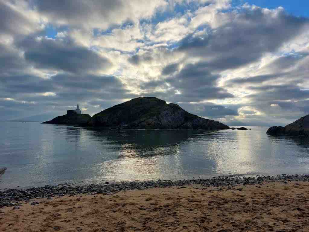 A serene coastal scene captures a rocky island under a cloudy sky. The water reflects the soft hues of the clouds, creating a calm ambiance. A sandy beach is visible in the foreground, while the distant island features a structure, adding depth to the landscape.