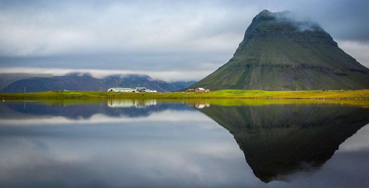 A serene landscape is captured, featuring a prominent mountain rising above a calm body of water. The reflection of the mountain on the water's surface creates a symmetrical view, while green fields and subtle clouds enhance the tranquil atmosphere of the surrounding area.