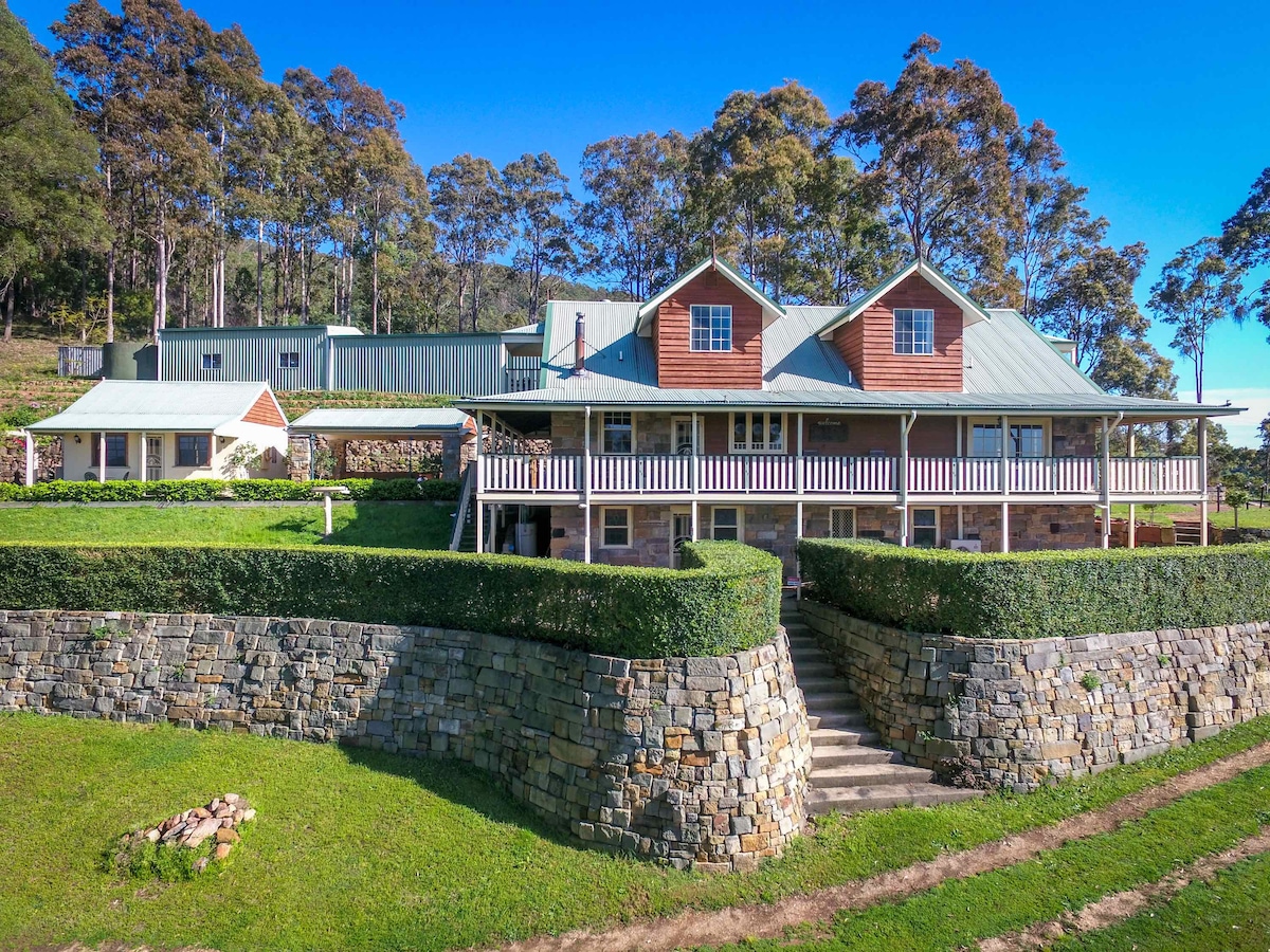 The outdoor view of Bimbadeen Mountain Retreat showcases a unique stone home with a welcoming porch. Surrounded by manicured greenery, the structure is elevated, providing a picturesque backdrop of trees and clear blue sky, creating an inviting atmosphere for relaxation.