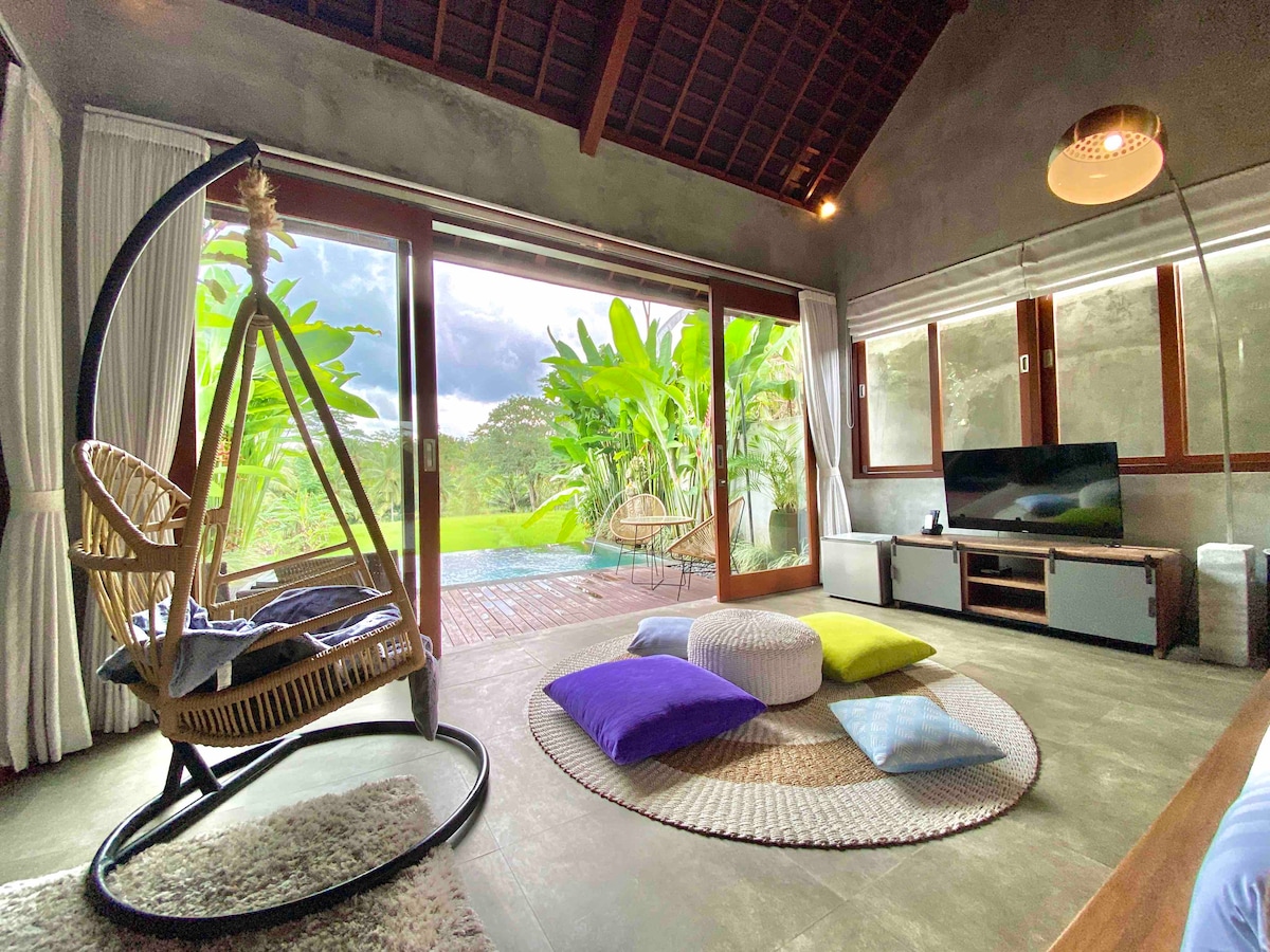 A spacious living area features large glass doors that open to a scenic view of lush greenery and an infinity pool. A hanging chair, colorful cushions, and a rug offer a cozy atmosphere. Natural light filters through the curtains, enhancing the serene environment.