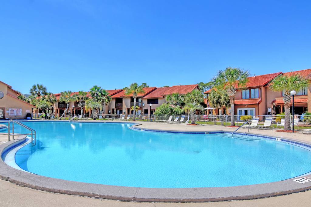 A large, circular swimming pool is set amidst lush palm trees and landscaped areas. Lounge chairs are arranged around the pool, which reflects clear blue skies above. The inviting pool area is bordered by a two-story townhouse complex with terracotta rooftops.