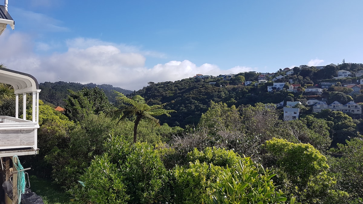 A scenic view of a lush, green landscape featuring hills and a variety of trees. In the foreground, diverse foliage adds depth to the image, while the rooftops of houses are visible in the distance against a clear blue sky.
