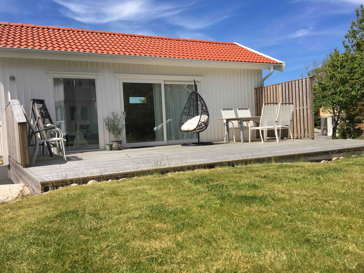 A newly built house features a spacious deck, surrounded by green grass. A hanging chair adds a touch of comfort, while outdoor seating is arranged for relaxation. Large glass doors allow natural light to fill the interior, enhancing the inviting outdoor space.