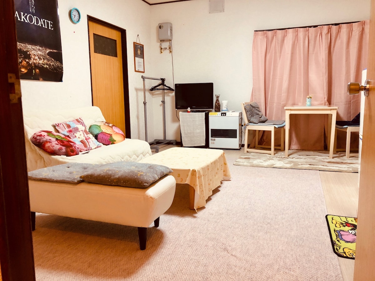 A living area features a white sofa positioned beside a low table, with colorful cushions adding comfort. A television and air conditioning unit are visible. Two chairs are arranged around a small dining table, with soft lighting casting a warm glow through the curtain-covered window.