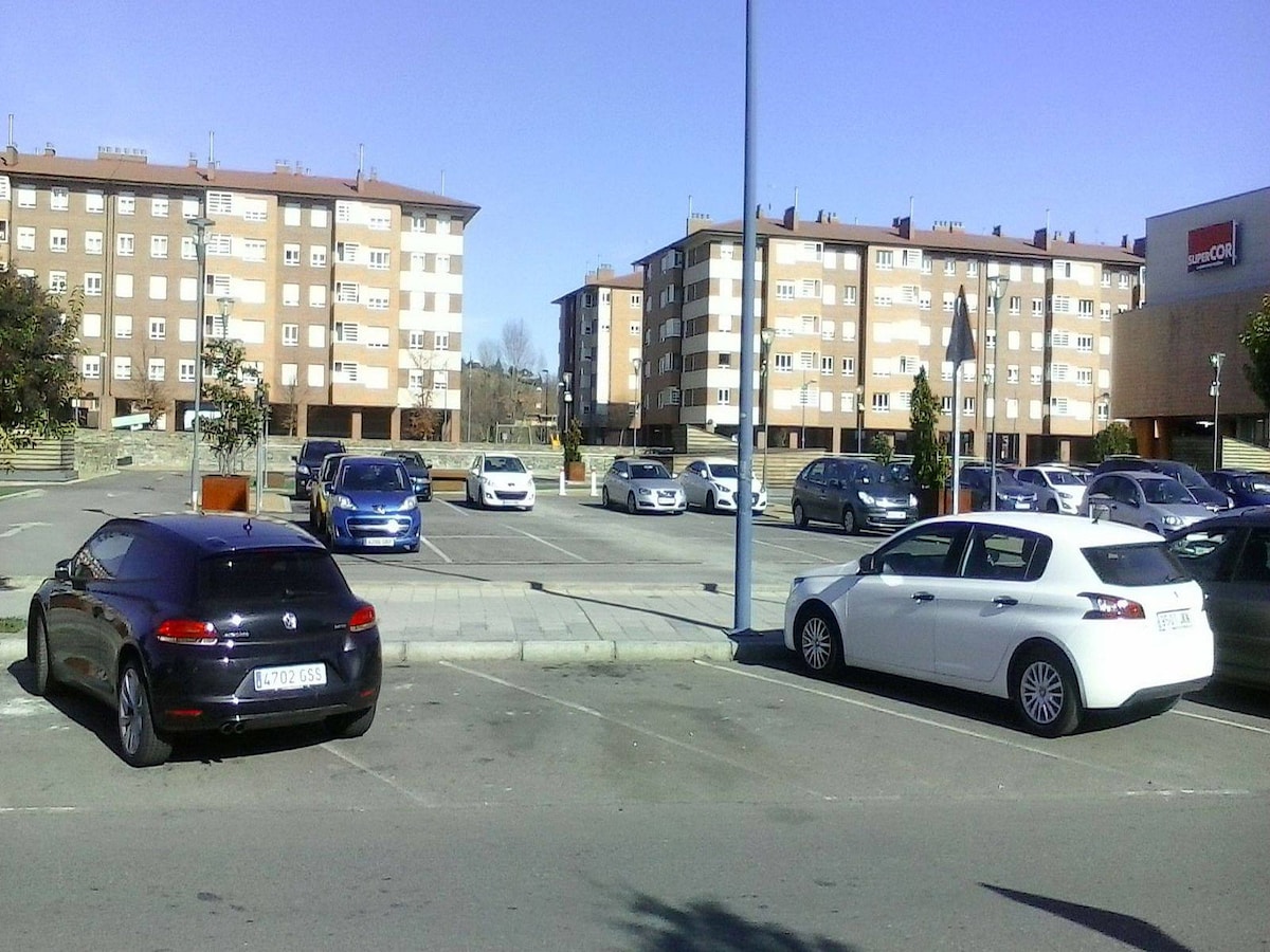 A residential parking lot is depicted, featuring several parked cars in varying colors, including black and white vehicles. Surrounding the lot are beige apartment buildings, contributing to the urban setting. Clear blue skies are visible above, indicating a sunny day.
