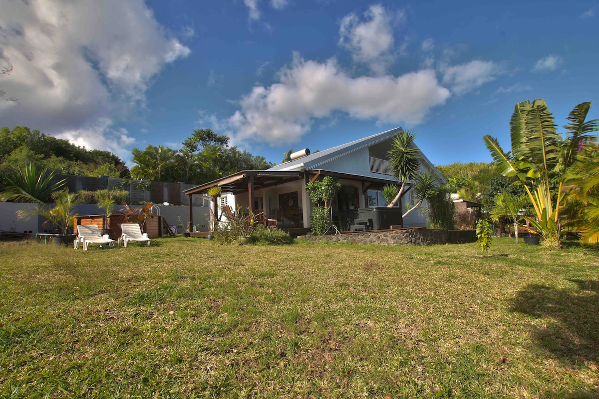 A spacious villa is surrounded by a lush garden, featuring ample green grass and various tropical plants. A covered terrace can be seen extending from the villa, providing shaded seating areas. The blue sky overhead is dotted with clouds, enhancing the inviting outdoor space.