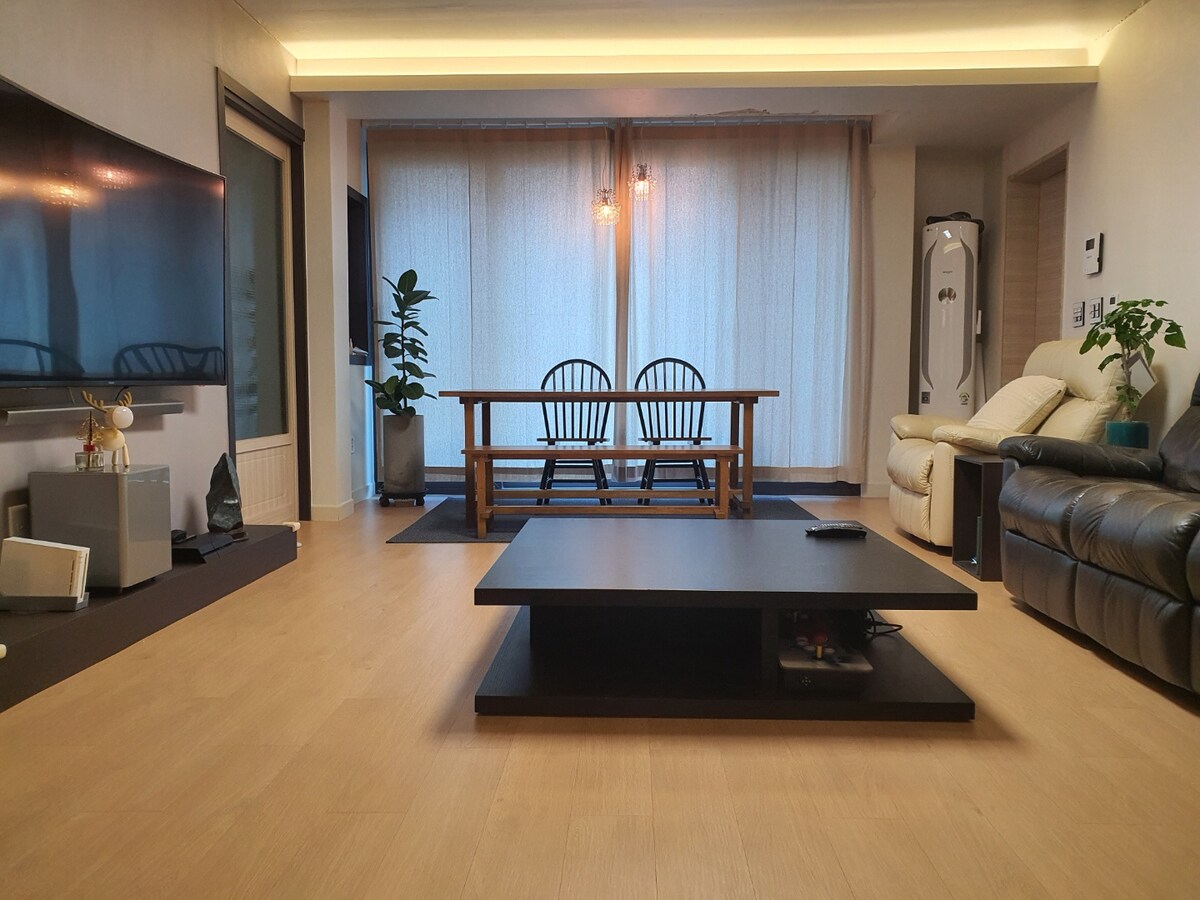 A spacious living area features a modern design with a low black coffee table centered in front of a comfortable reclining sofa. A wooden dining table with matching chairs is positioned by large windows, adorned with sheer curtains that allow natural light to filter in.