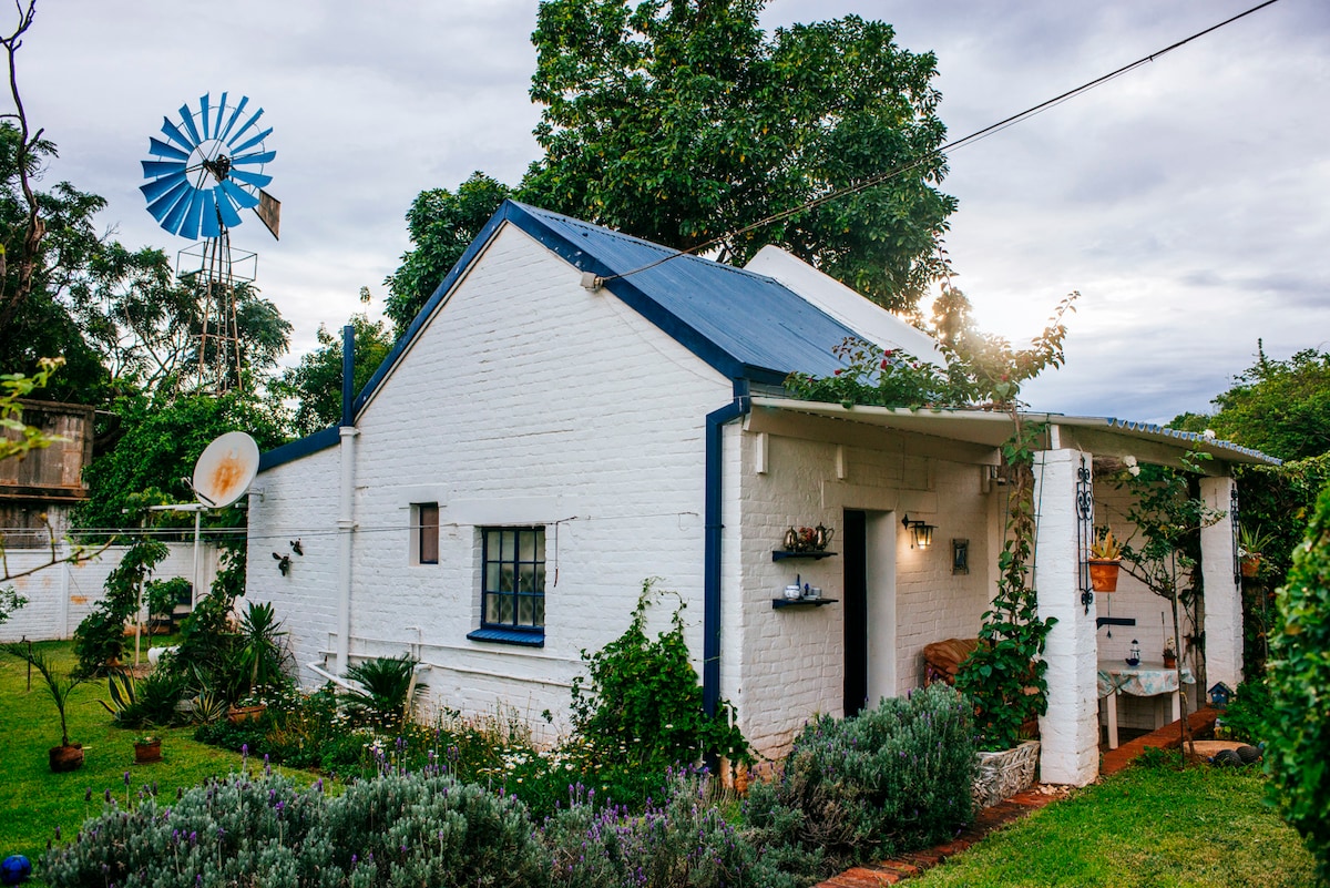 A charming Greek-themed cottage is surrounded by greenery, featuring a white exterior with blue accents. A windmill stands nearby, while the garden is adorned with various plants. A shaded porch area invites relaxation, blending indoor and outdoor living spaces.