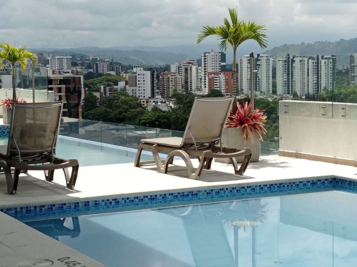 An outdoor pool area is highlighted, featuring lounge chairs and surrounding potted plants. The clear water reflects the skyline of the city, with modern buildings and greenery in the distance against a backdrop of cloudy skies.