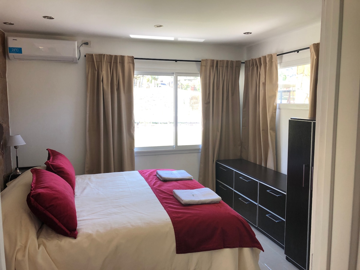 A bright bedroom features a spacious bed with red accent pillows and neatly arranged towels. Natural light enters through large windows adorned with light curtains. A dresser stands against the wall, providing ample storage space, while a small lamp adds functionality.