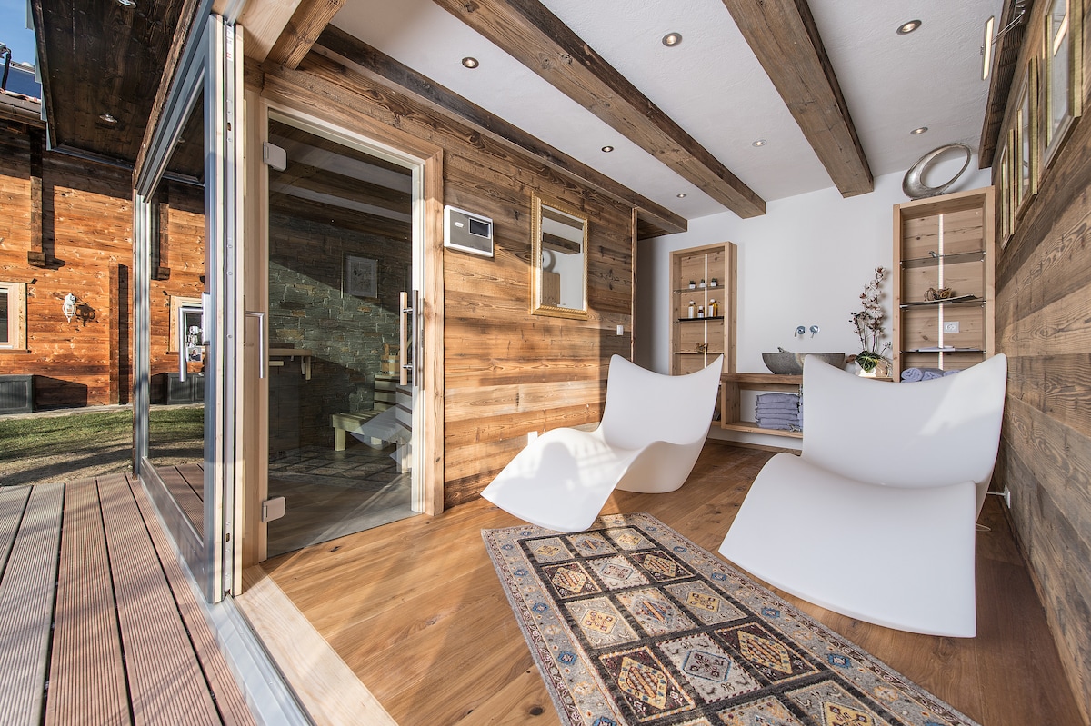 A modern lounge area features two white lounge chairs positioned on warm wooden flooring. Large glass doors provide access to an outdoor space, while wooden beams add character to the rustic ambiance. A patterned area rug enhances the cozy atmosphere.