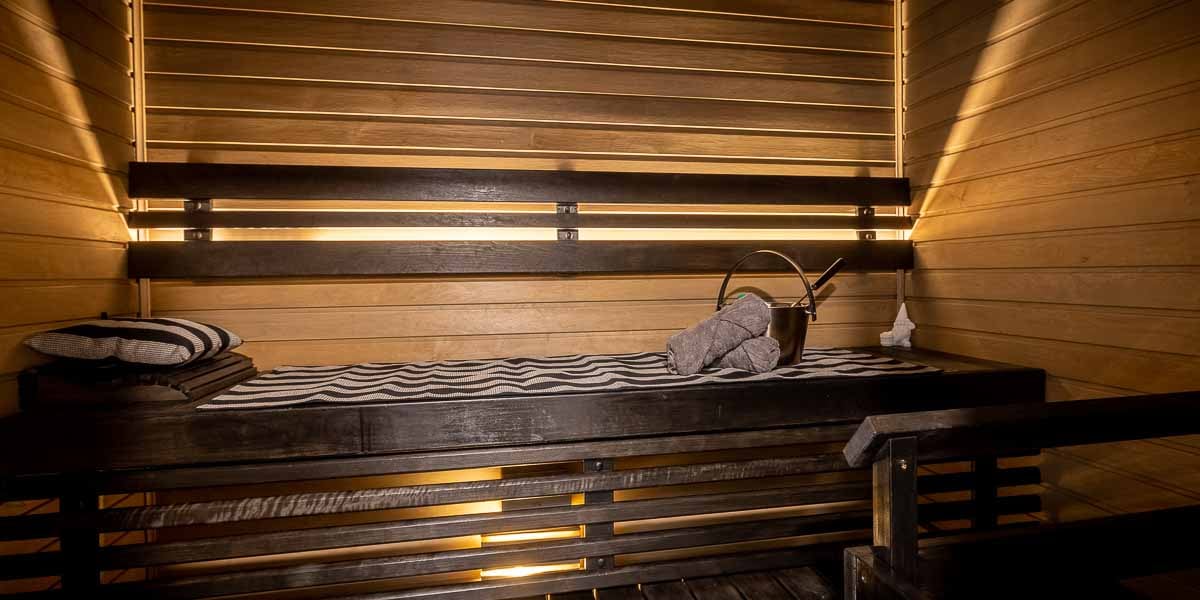 A wooden sauna interior features built-in benches and subtle recessed lighting. Soft, patterned cushions are placed on the benches, while a folded towel and a wooden bucket are positioned nearby, creating a calming and warm atmosphere.