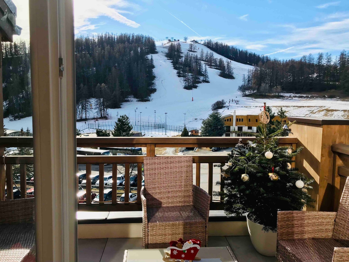A balcony is furnished with two wicker chairs and a table, overlooking snow-covered slopes and trees. A decorated Christmas tree adds a festive touch, while the clear blue sky enhances the serene mountain views.