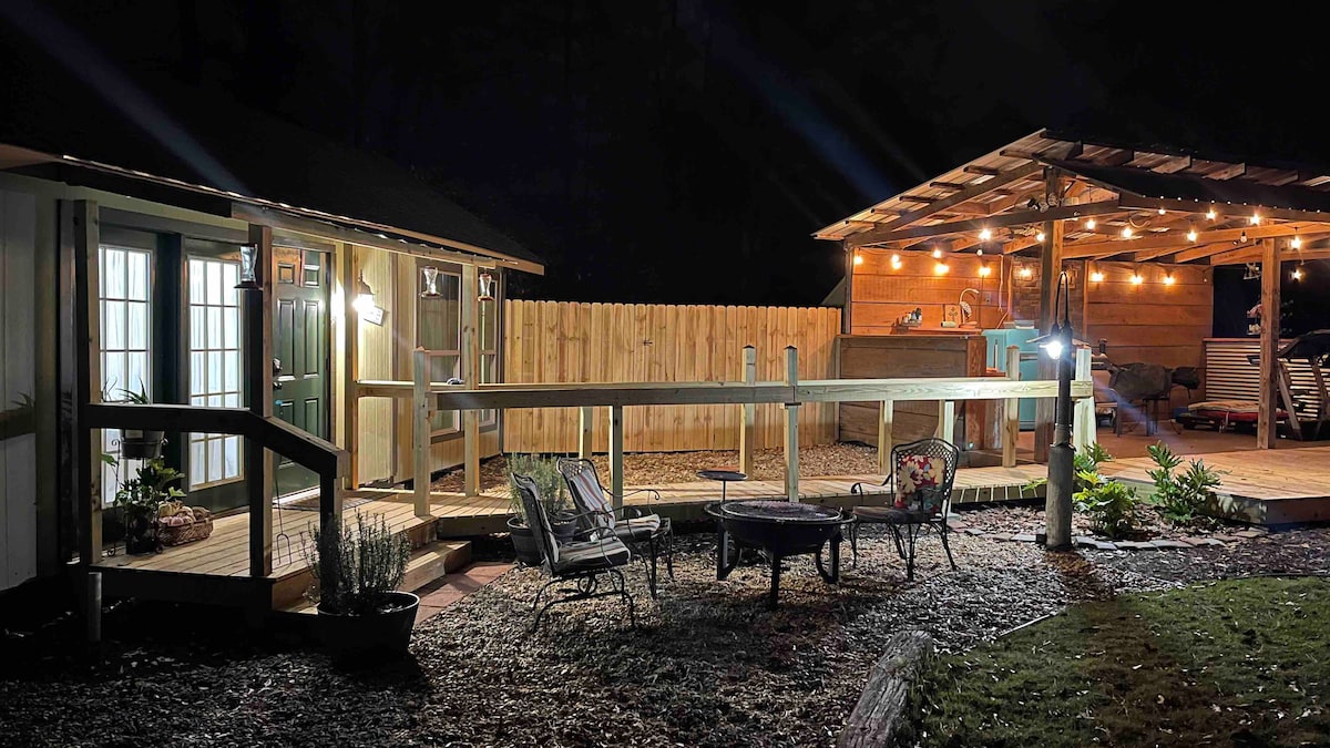 The outdoor area is illuminated under a night sky, featuring a cozy fire pit surrounded by chairs. A spacious wooden deck with warm lighting is visible, along with a partially covered outdoor kitchen area. The tiny house entrance is framed by a welcoming green door.