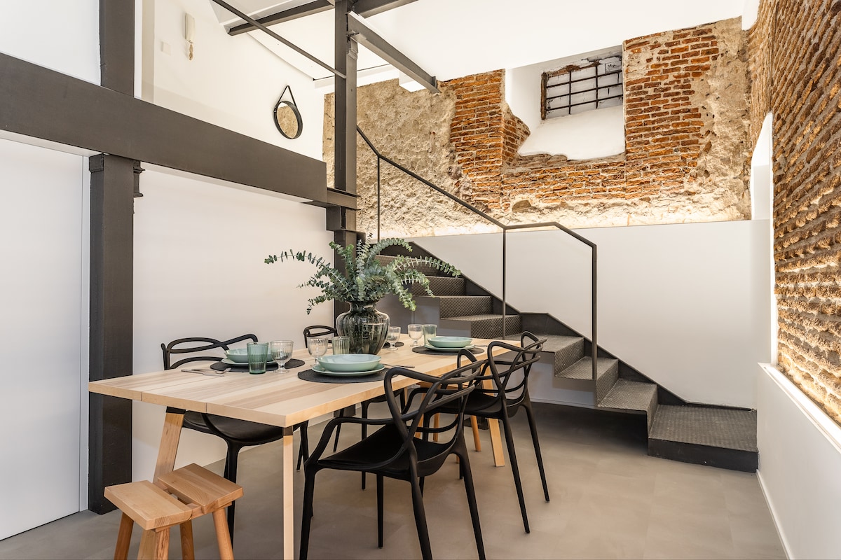 A spacious dining area features a wooden table set for six, accompanied by black chairs. The wall showcases exposed brick and stone, adding texture to the modern setting. A plant centerpieces the table, while a staircase leads to an upper level.