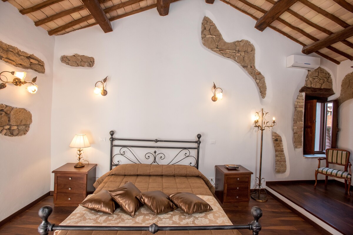 A spacious bedroom features a decorative metal bed frame with an earthy-toned bedding set. Side tables with lamps are positioned on either side. Exposed stone accents line the walls, and a cozy chair is situated near a window, allowing natural light to enter the room.