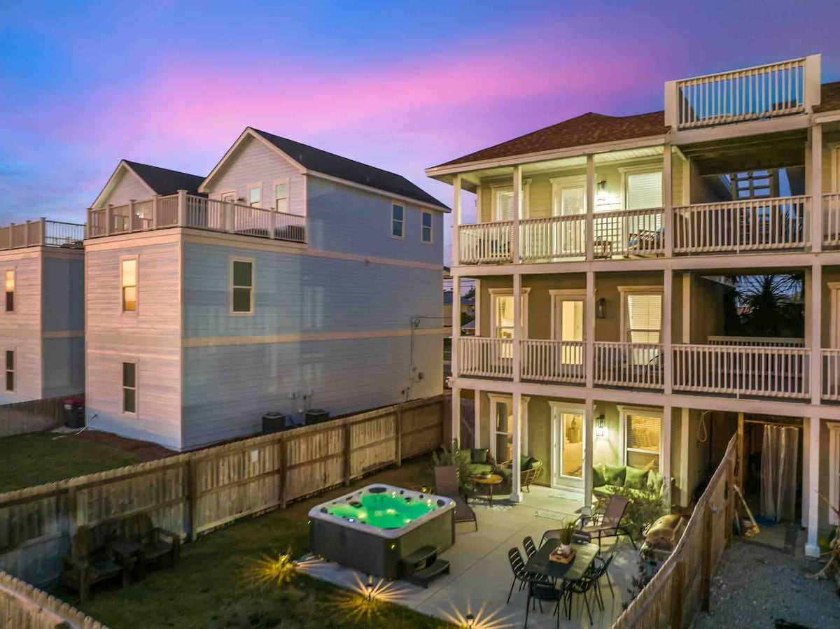 A modern beach house is viewed at dusk, featuring three stories with balconies. A private hot tub sits on the patio, surrounded by outdoor seating. The surrounding yard is enclosed by a wooden fence, providing a serene atmosphere for relaxation.