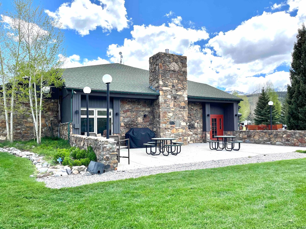 A stone-clad clubhouse is set against a backdrop of blue sky with fluffy clouds. The patio features picnic tables and a grill, surrounded by well-maintained grass and landscaping. Tall trees provide shade, enhancing the outdoor recreation area.