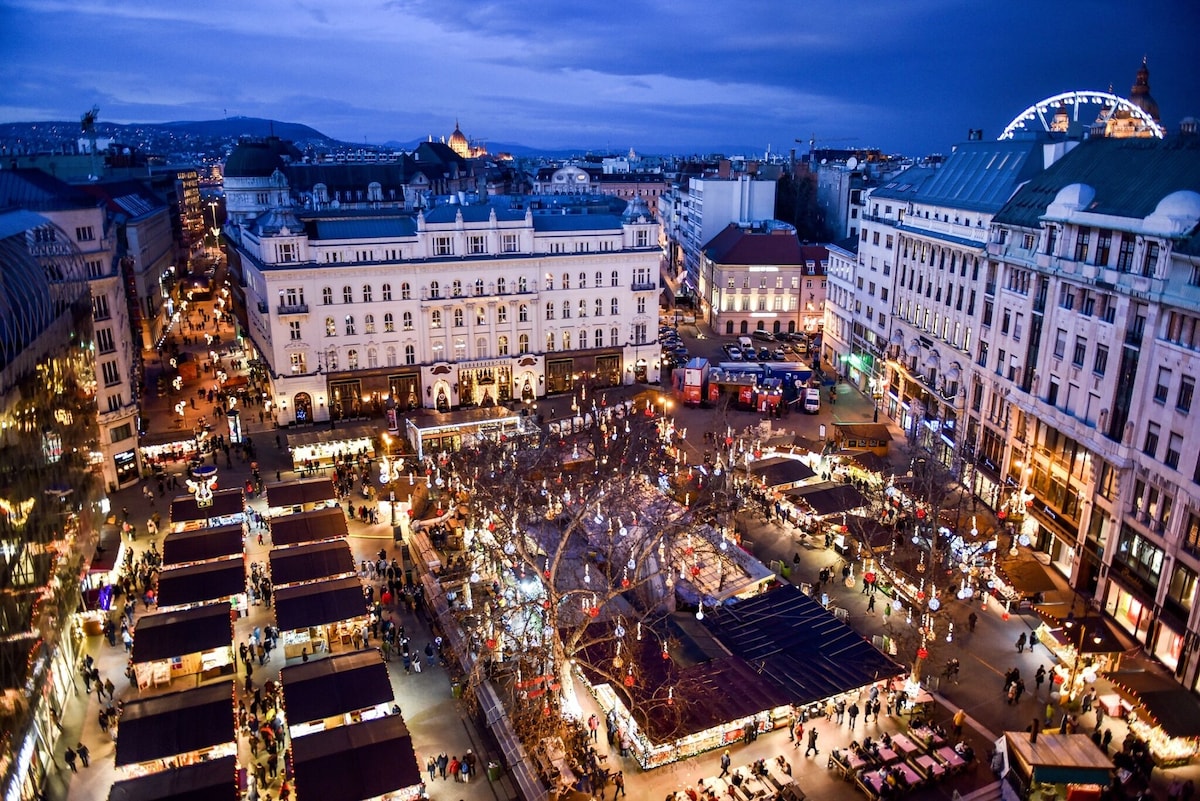 An aerial view captures a bustling city square adorned with market stalls in the evening. The vibrant atmosphere is highlighted by twinkling lights, with a backdrop of historic buildings and a Ferris wheel in the distance, suggesting a lively urban scene.